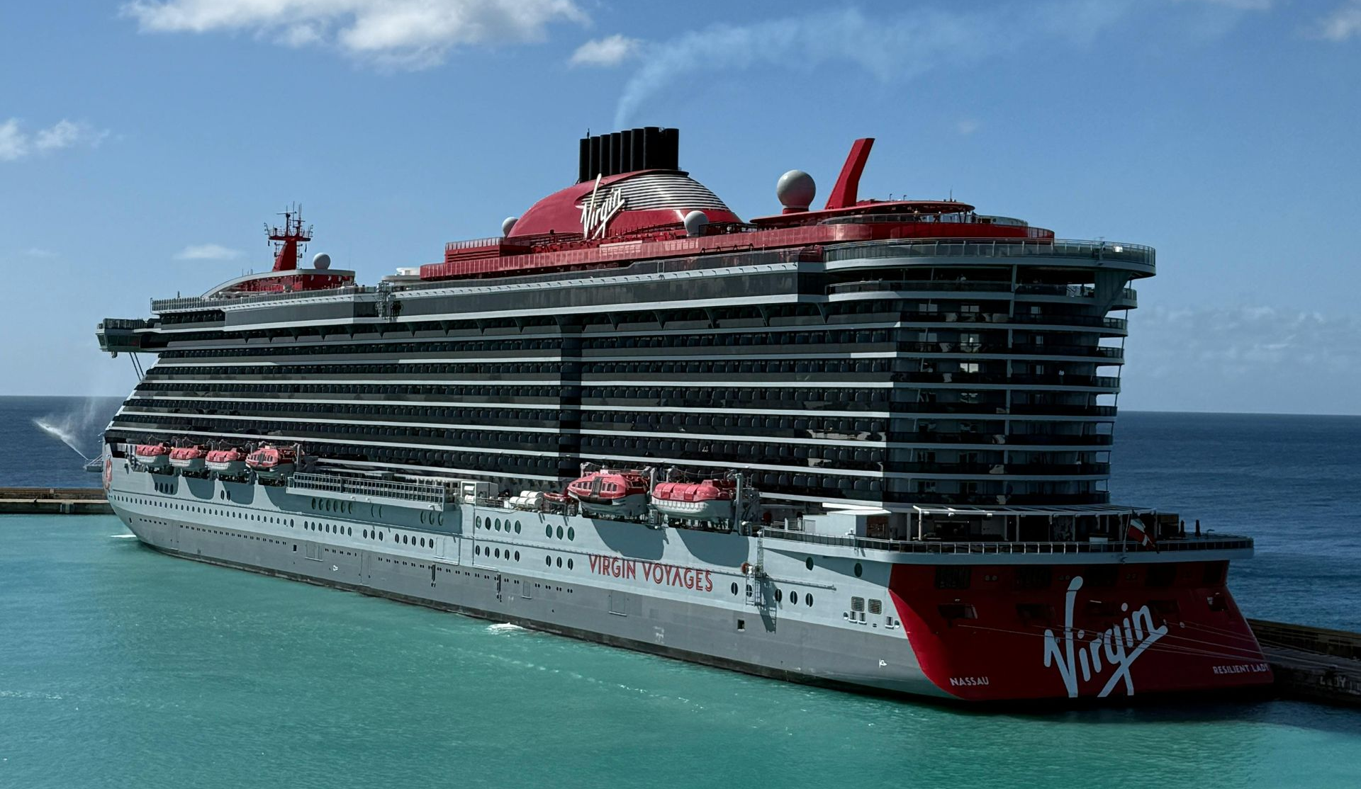 Large gray and red Virgin Voyages cruise ship in turquoise water, docked near a breakwater on a sunny day.