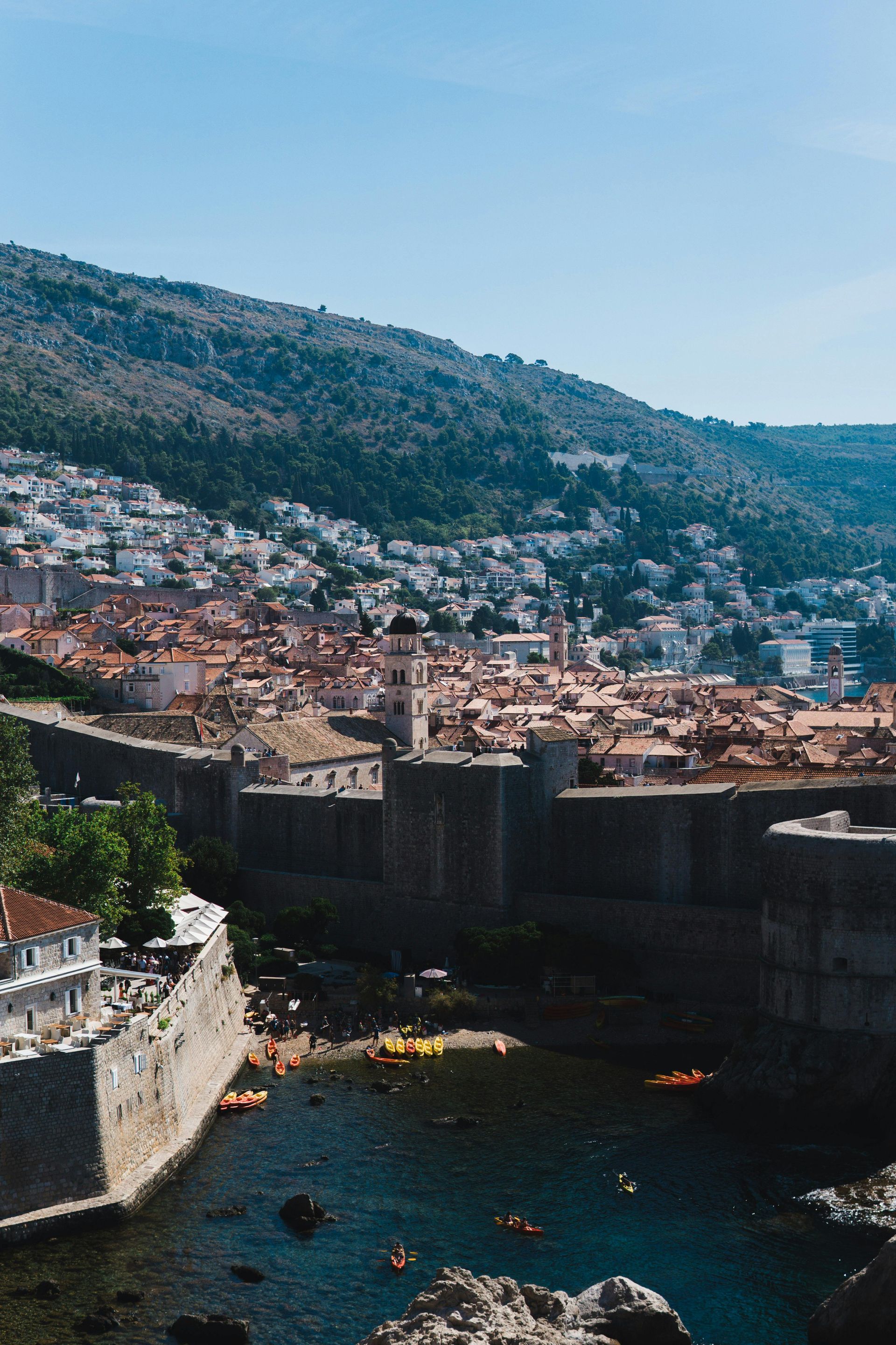 View of Dubrovnik's old town and walls along the coast with a mountainous backdrop under a bright sky.