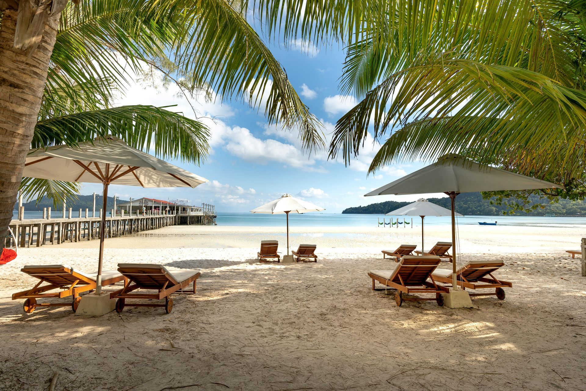 Beach scene with lounge chairs, umbrellas, palm trees, and a pier.
