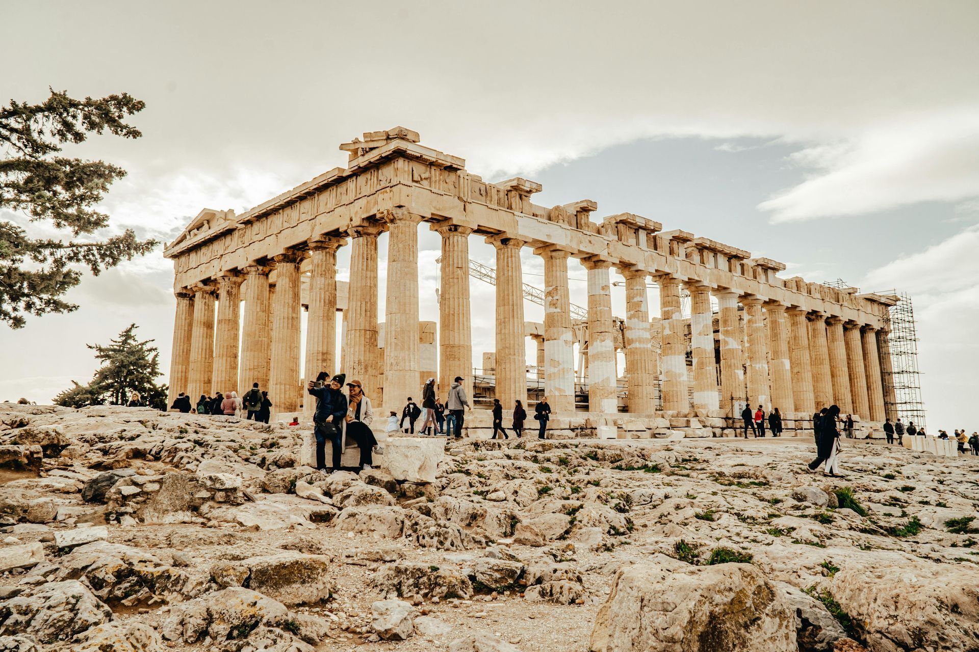 Parthenon temple in Athens with tourists on the rocky ground below, under a cloudy sky.