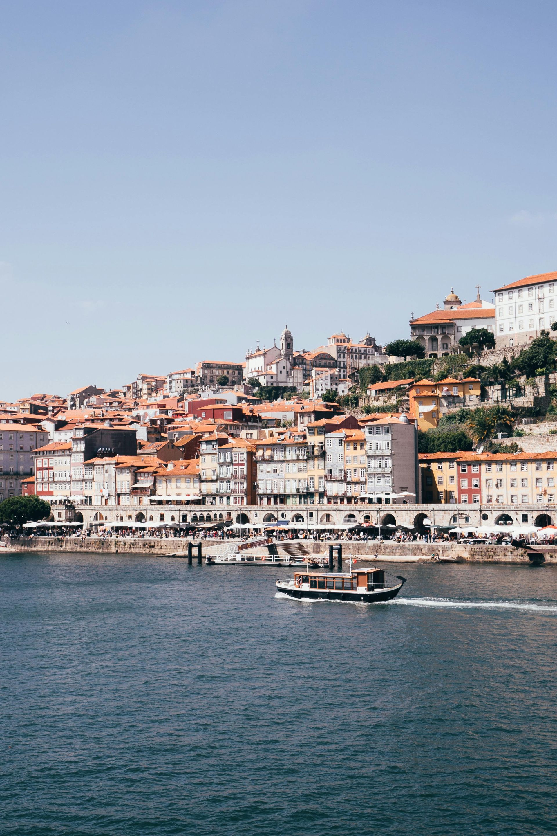 Boat sails on blue water in front of Porto, Portugal, with colorful buildings and clear sky.