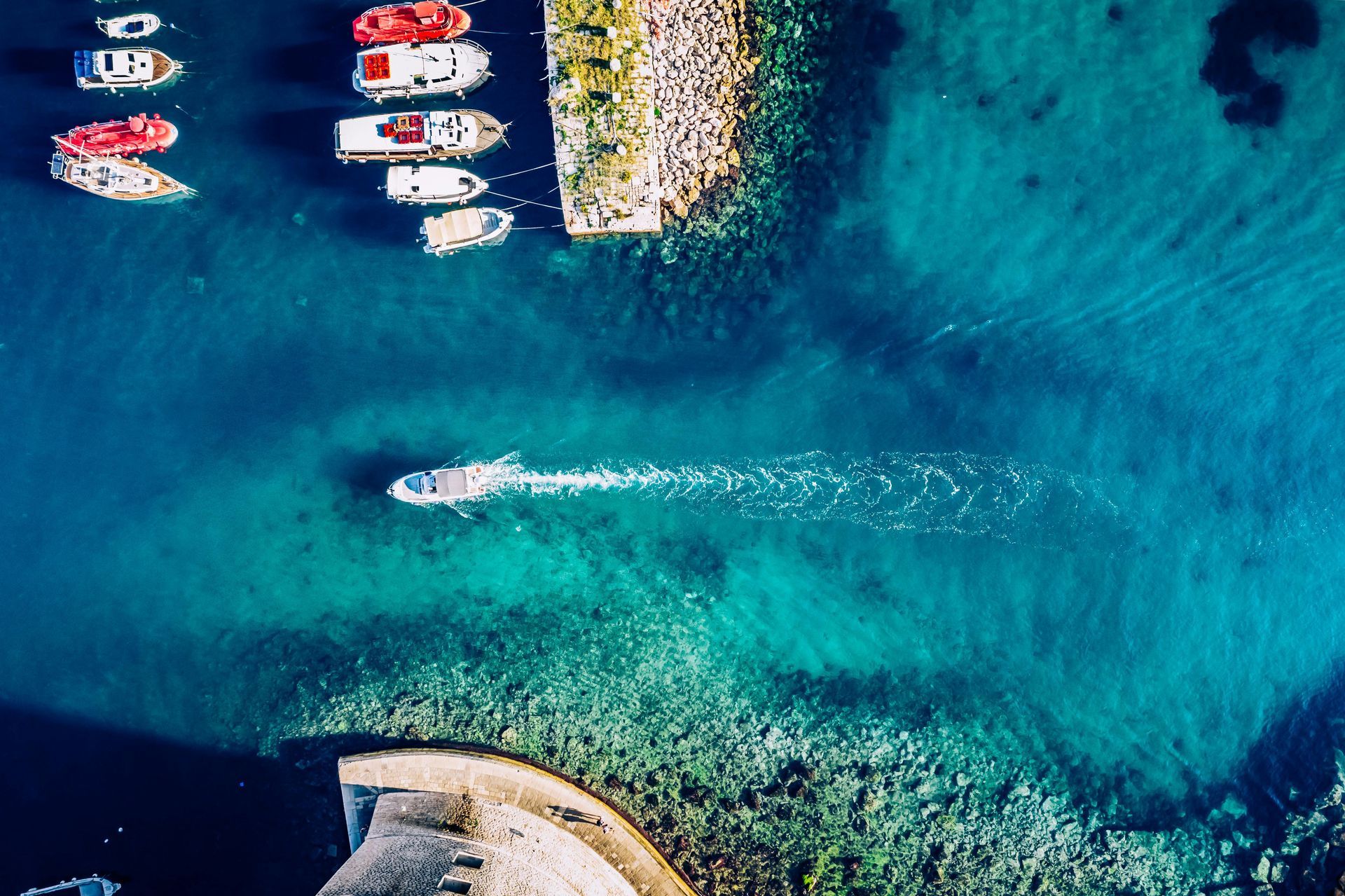 Aerial view of a boat leaving a harbor of clear, turquoise water, boats docked, and a stone wall.