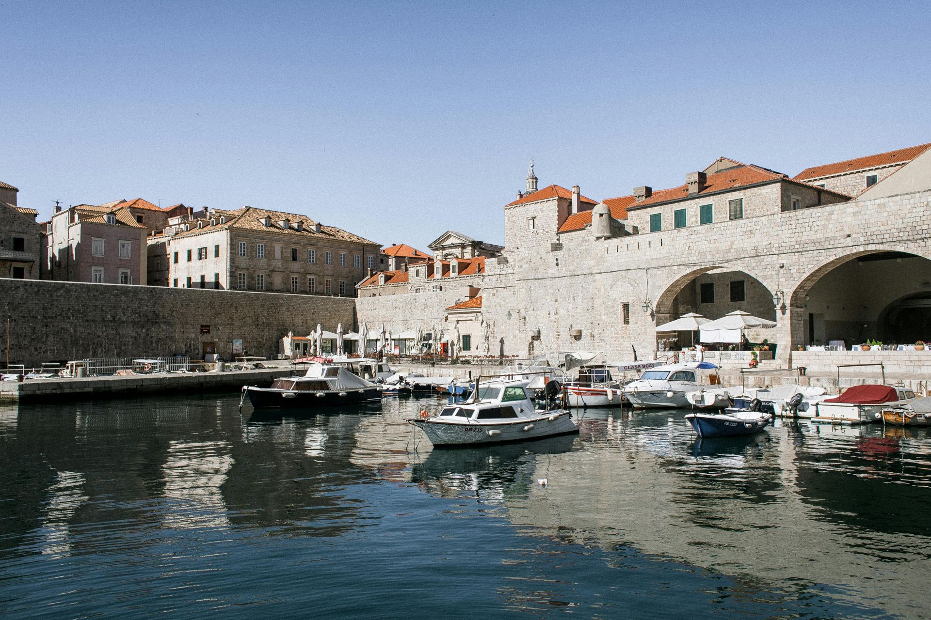 Boats docked in a harbor, old stone buildings line the water under a clear blue sky.