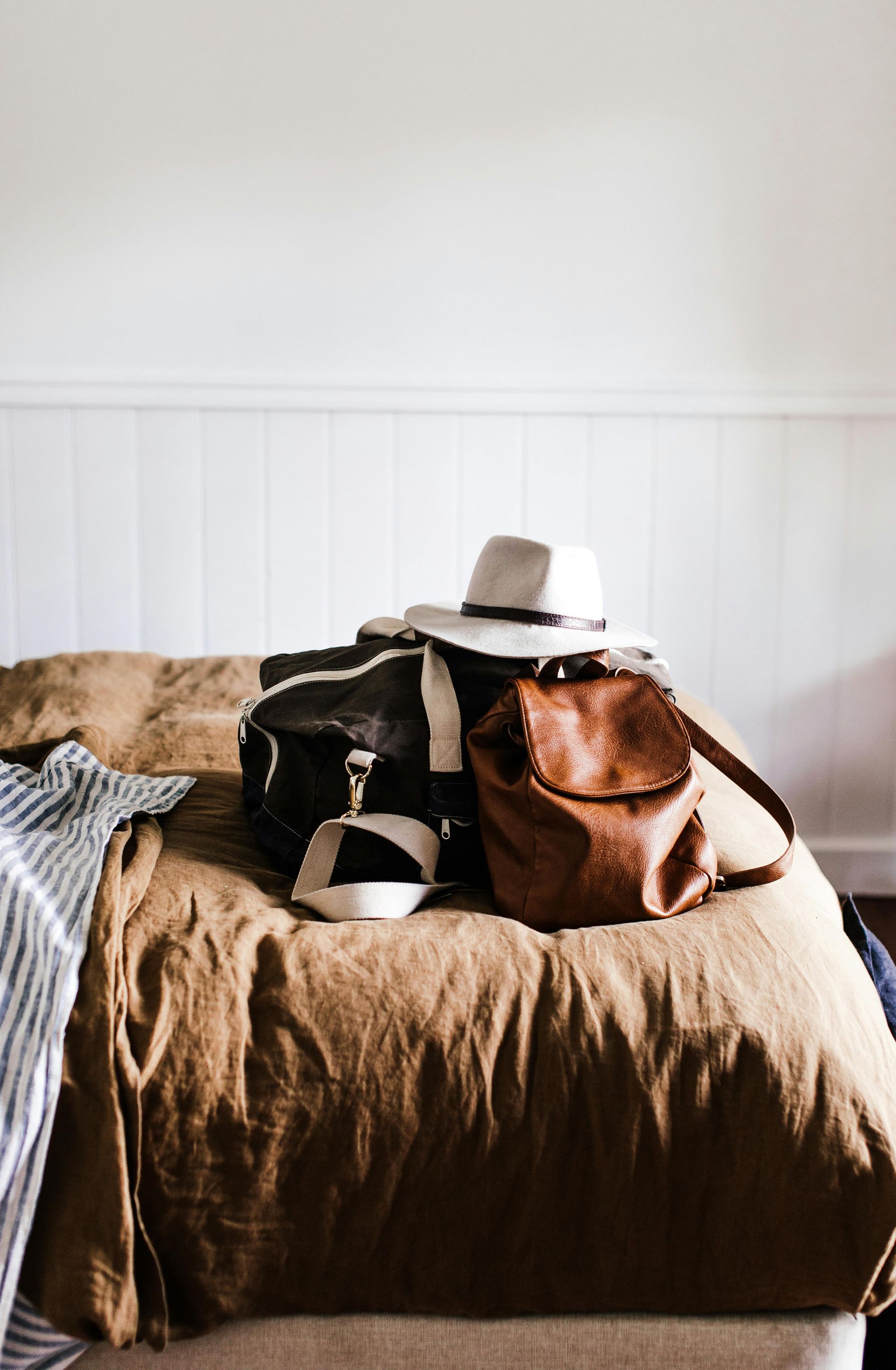 Bags, hat, and sandals on a bed with brown bedding, against a white paneled wall.