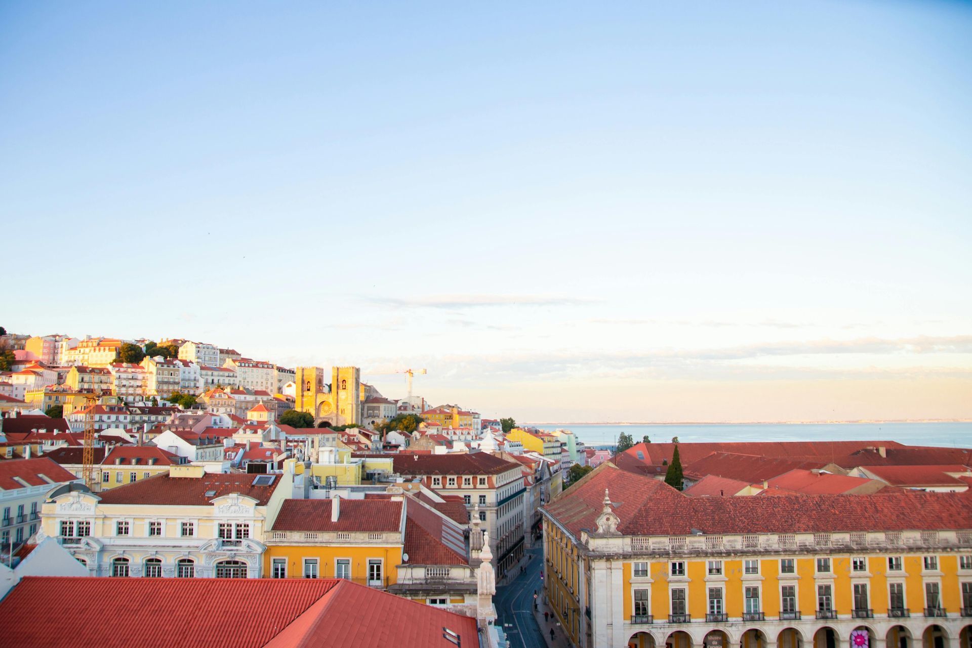 Rooftops and buildings of Lisbon, Portugal under a light blue sky, with a glimpse of the ocean.