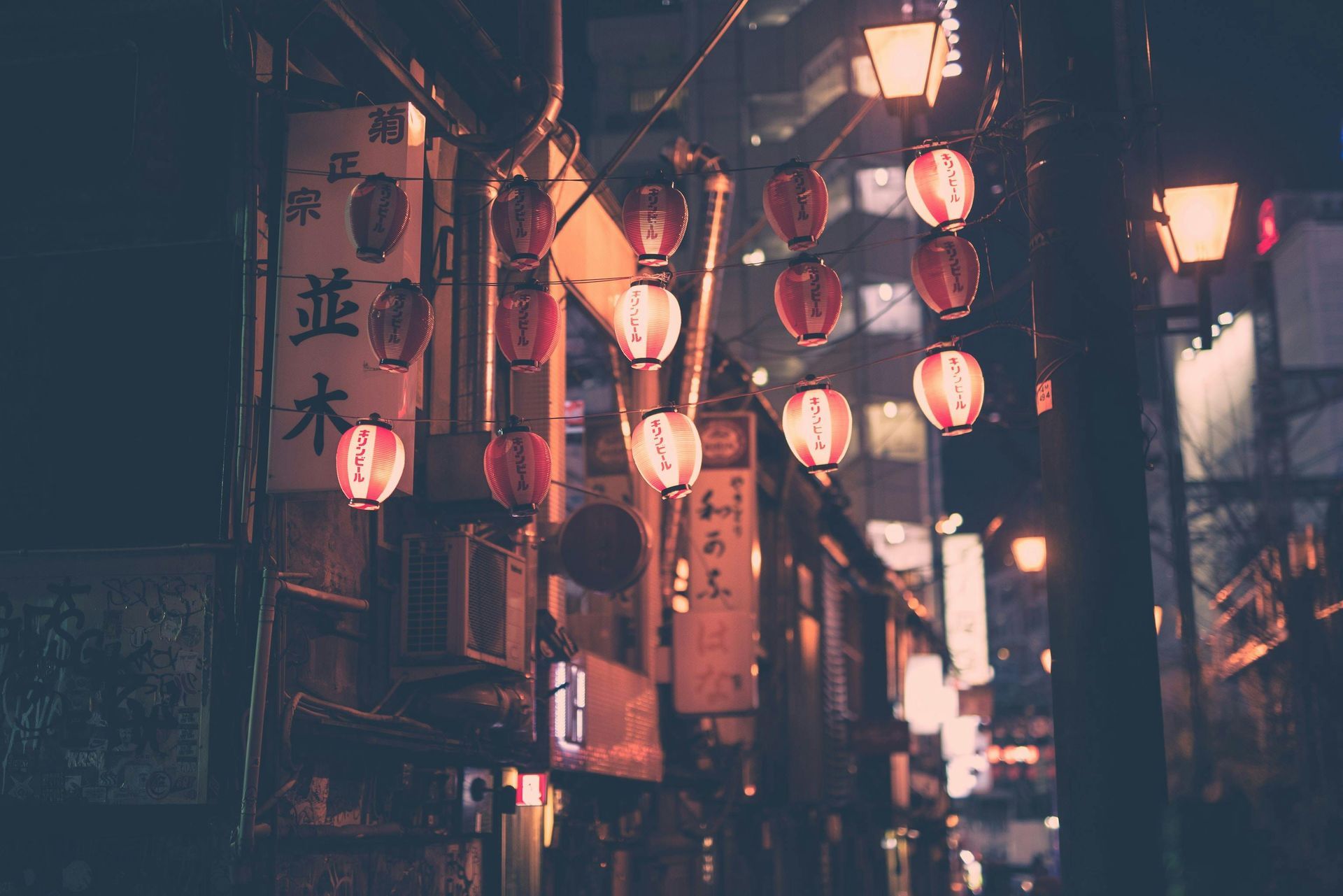 Night scene in a Japanese alley with red lanterns, shop signs, and street lights.