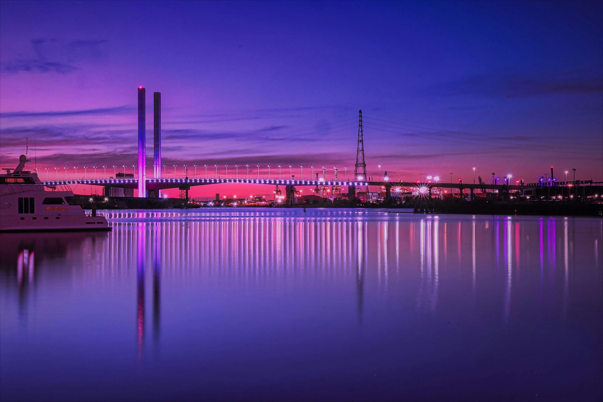 Night view of Melbourne's Bolte Bridge, lit with purple and pink lights, reflected in the still water.