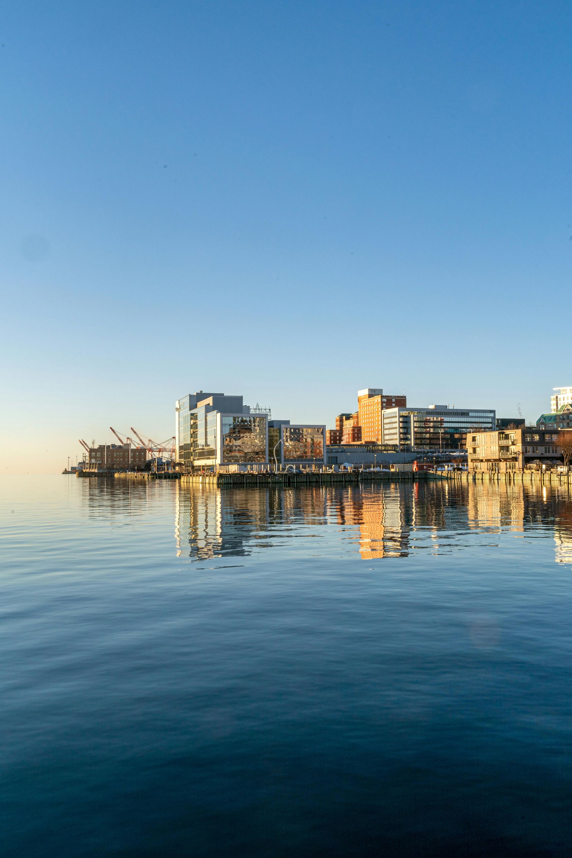 City buildings reflecting on calm water under a clear blue sky.