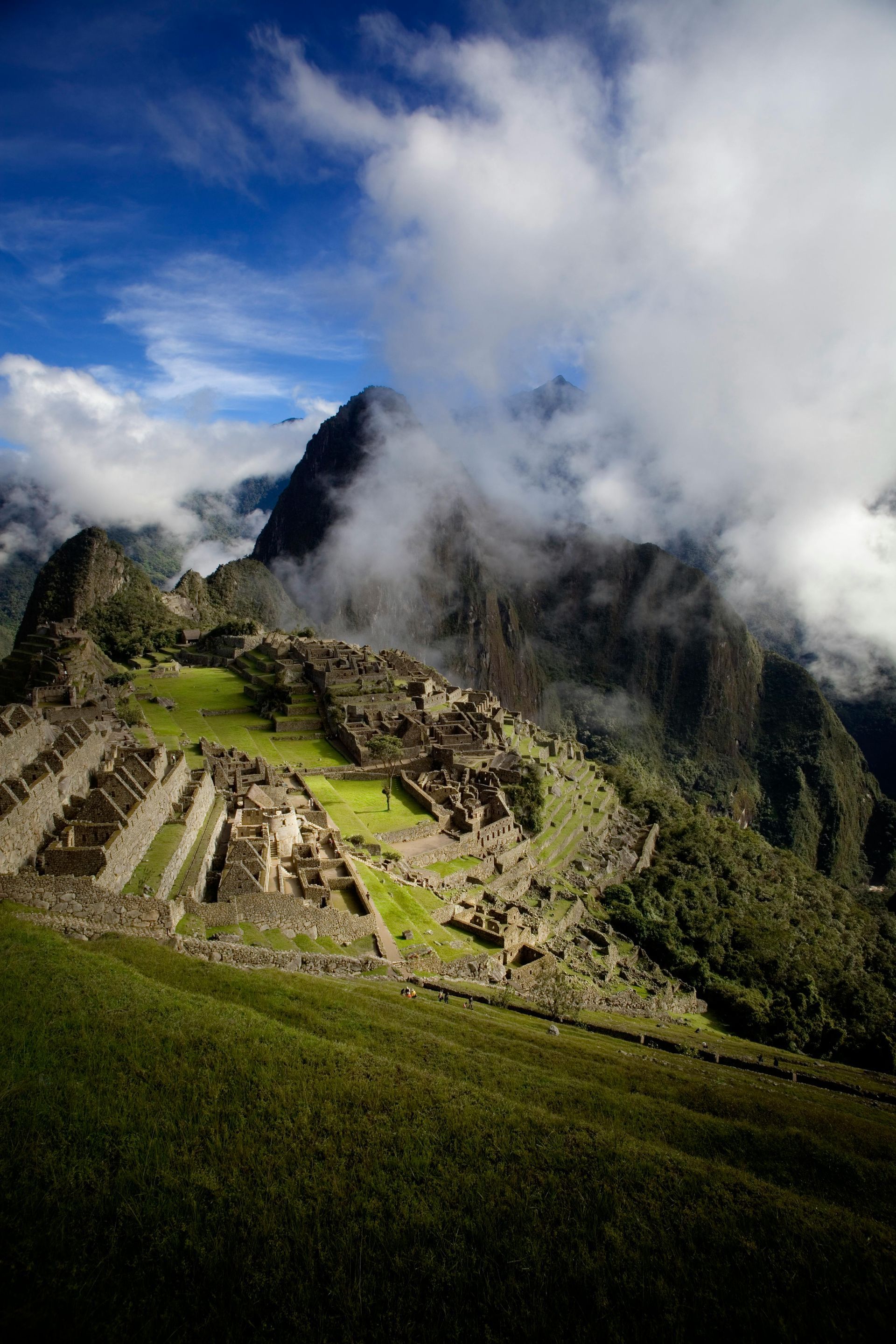 Machu Picchu ruins nestled in green mountains, partially shrouded in clouds under a blue sky.