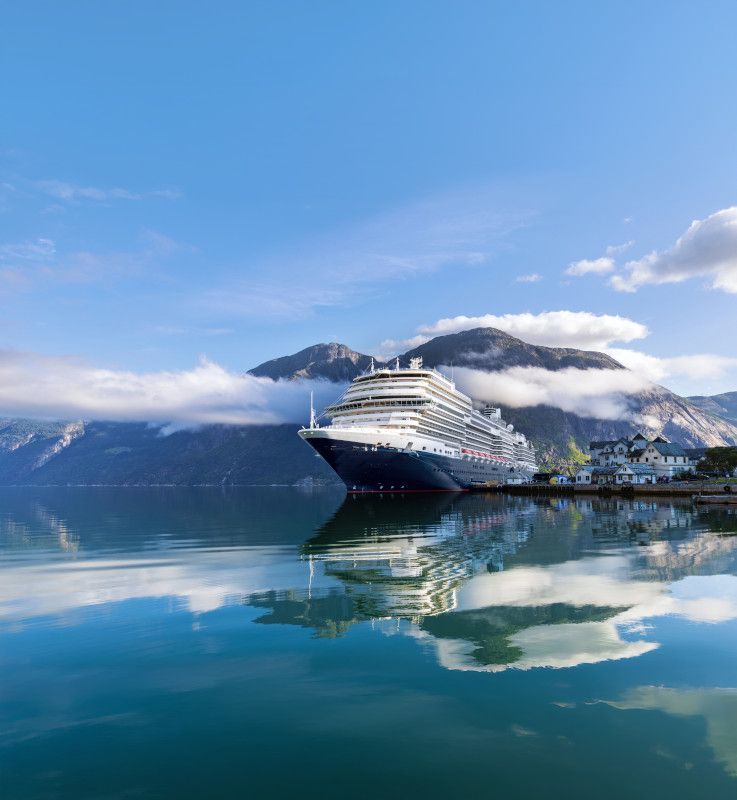 Cruise ship docked in a fjord, mountains in the background, blue water and sky, white clouds.