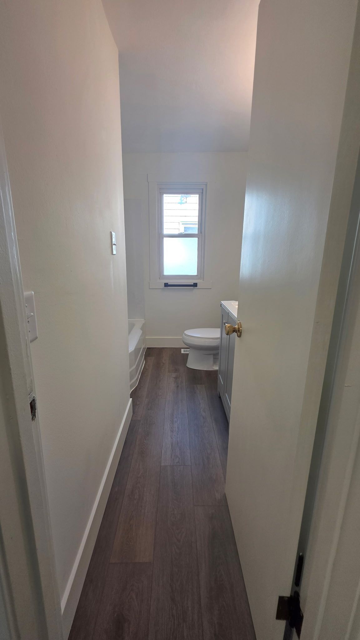 Narrow bathroom with a toilet, window, and wood-look flooring. White walls and door.