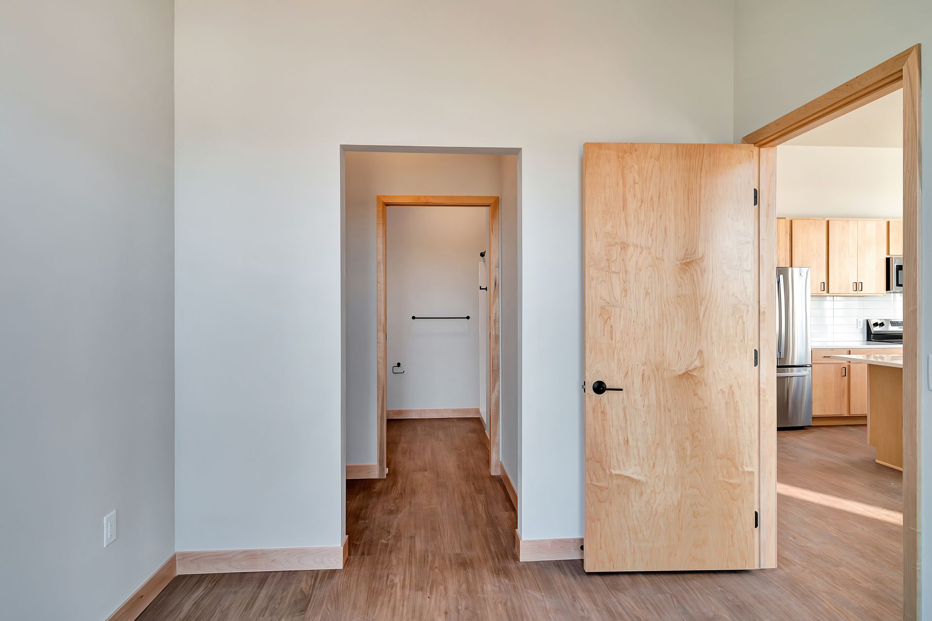 An empty room with a wooden door and a kitchen in the background.