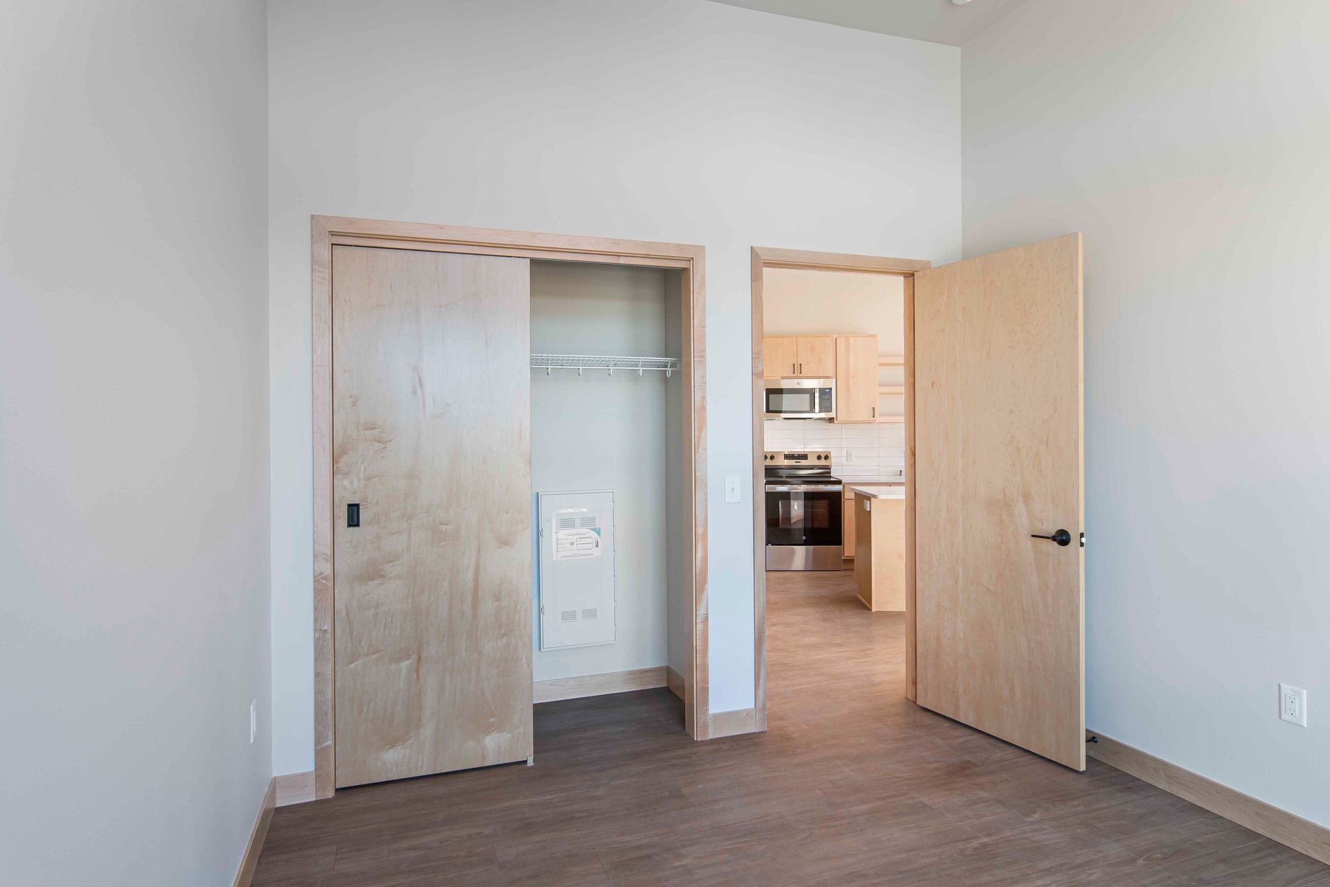 An empty bedroom with a closet and a kitchen in the background.