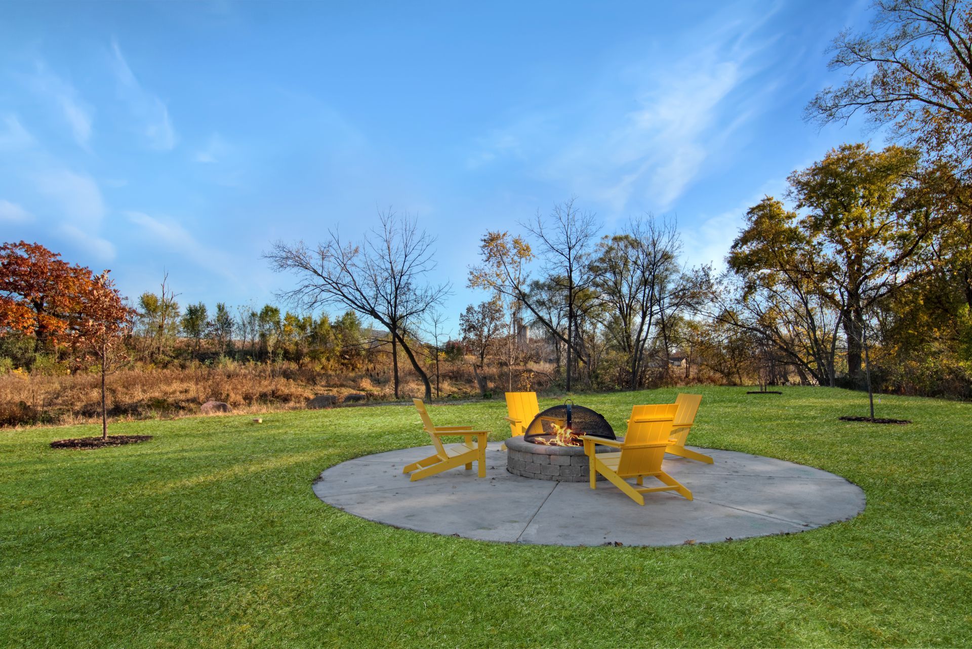 A fire pit in the middle of a lush green field with yellow chairs around it.