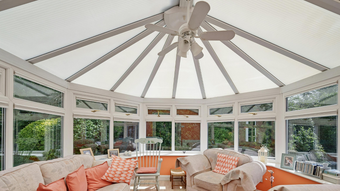 Sunroom with white frame and ceiling, beige couches, orange pillows, and view of greenery.