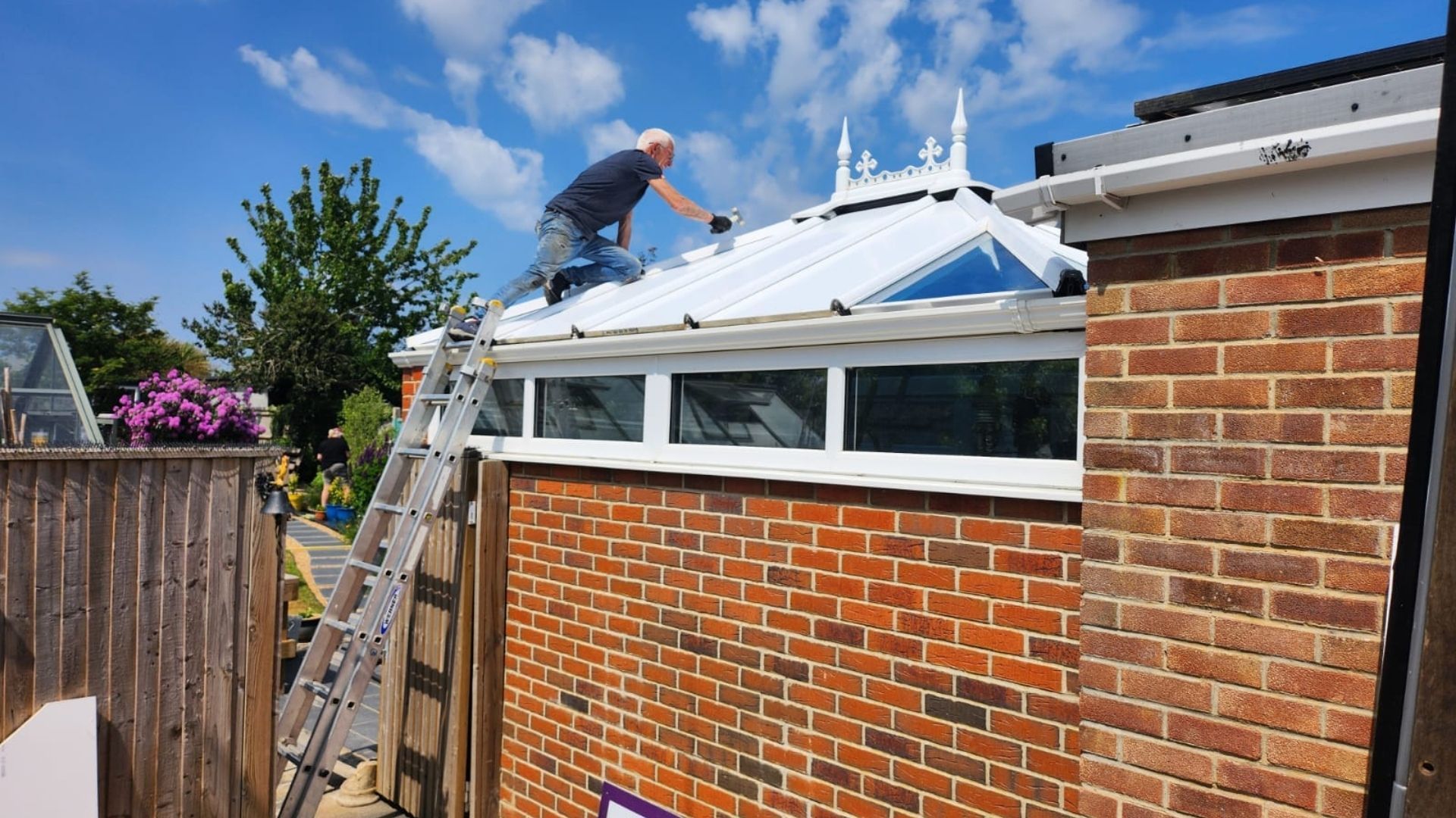 Man painting a white roof on a brick building, standing on the roof, with a ladder and blue sky.