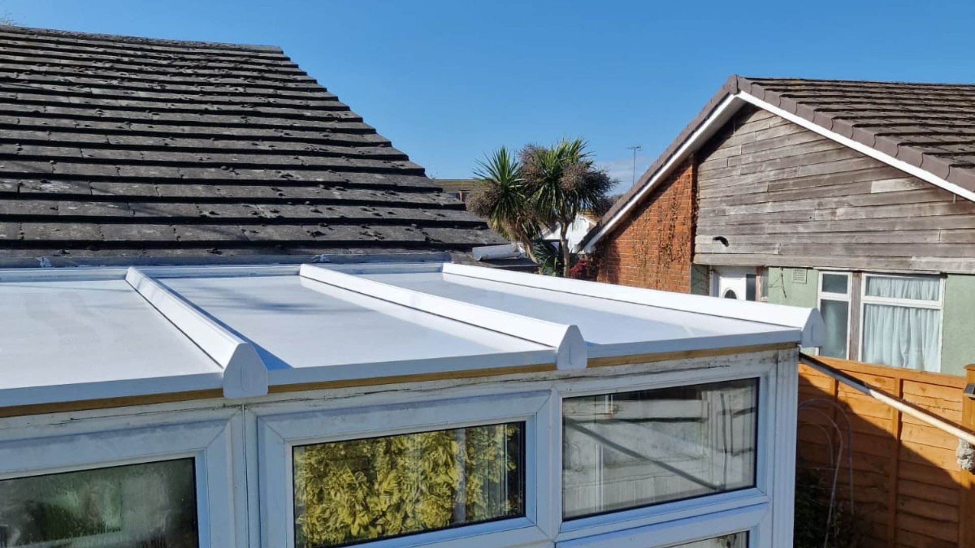 White-roofed conservatory with white window frames, next to a tiled roof and other buildings on a sunny day.