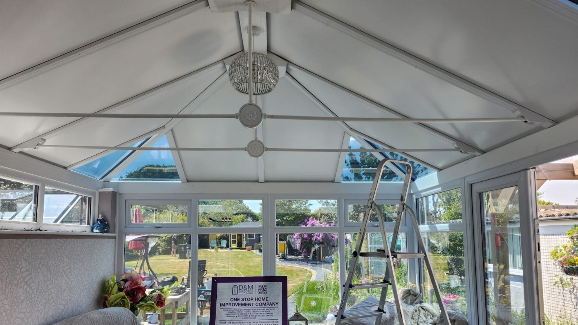 Interior view of a white sunroom with glass walls and ceiling. A ladder is present, along with a decorative light fixture.