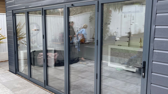 Gray-framed glass doors of a building, reflecting a couple inside. Exterior is gray siding and patio.