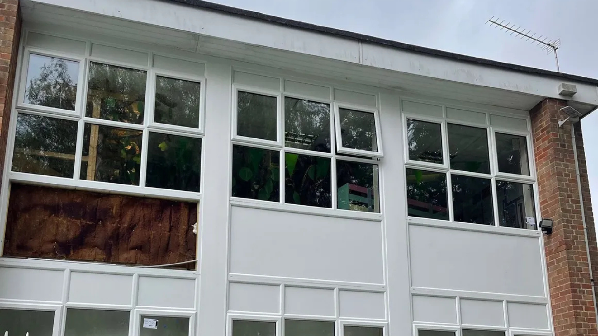 White-framed windows on a building. One window is covered. Exterior shot with greenery visible through the glass.