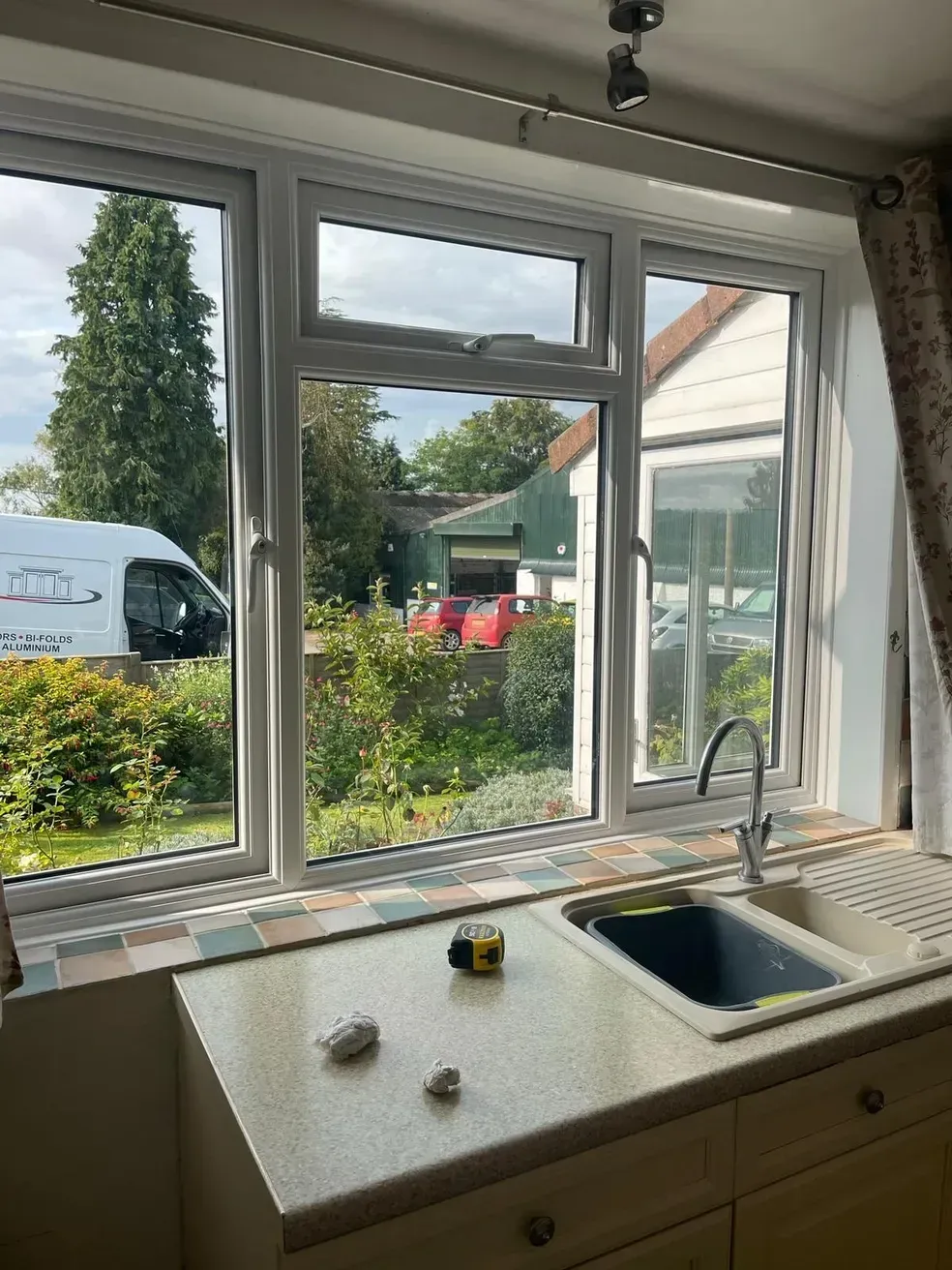 Kitchen window with a view of a garden and parked vehicles; a sink and counter are in the foreground.
