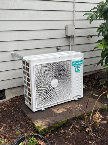 White outdoor air conditioner unit with a fan grille beside a gray house wall.