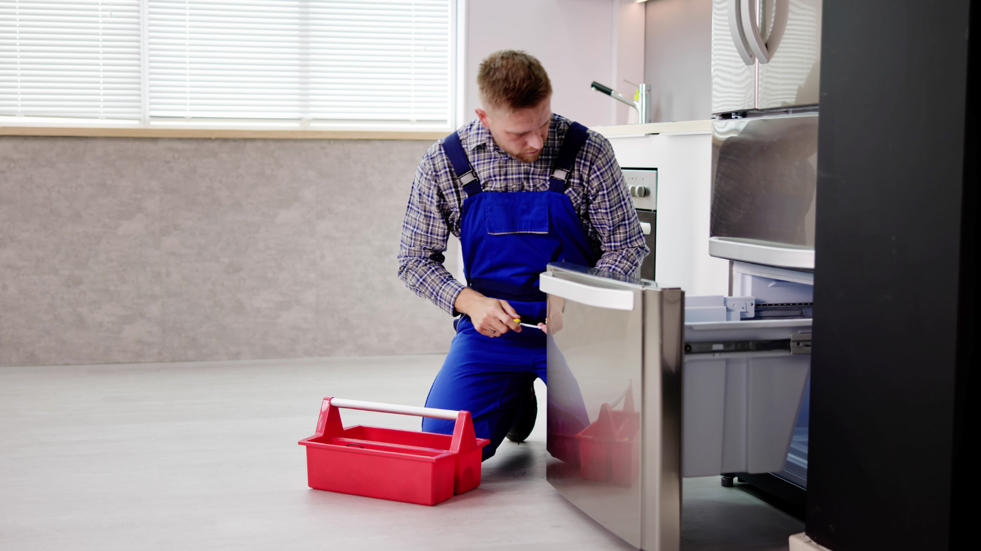 A man is kneeling on the floor fixing a refrigerator.