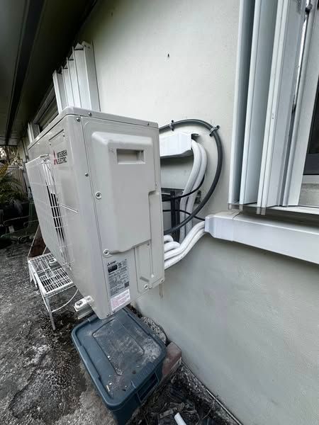 An air conditioning unit mounted on the side of a building, with pipes and cables running to the exterior. Grey and white colors dominate.