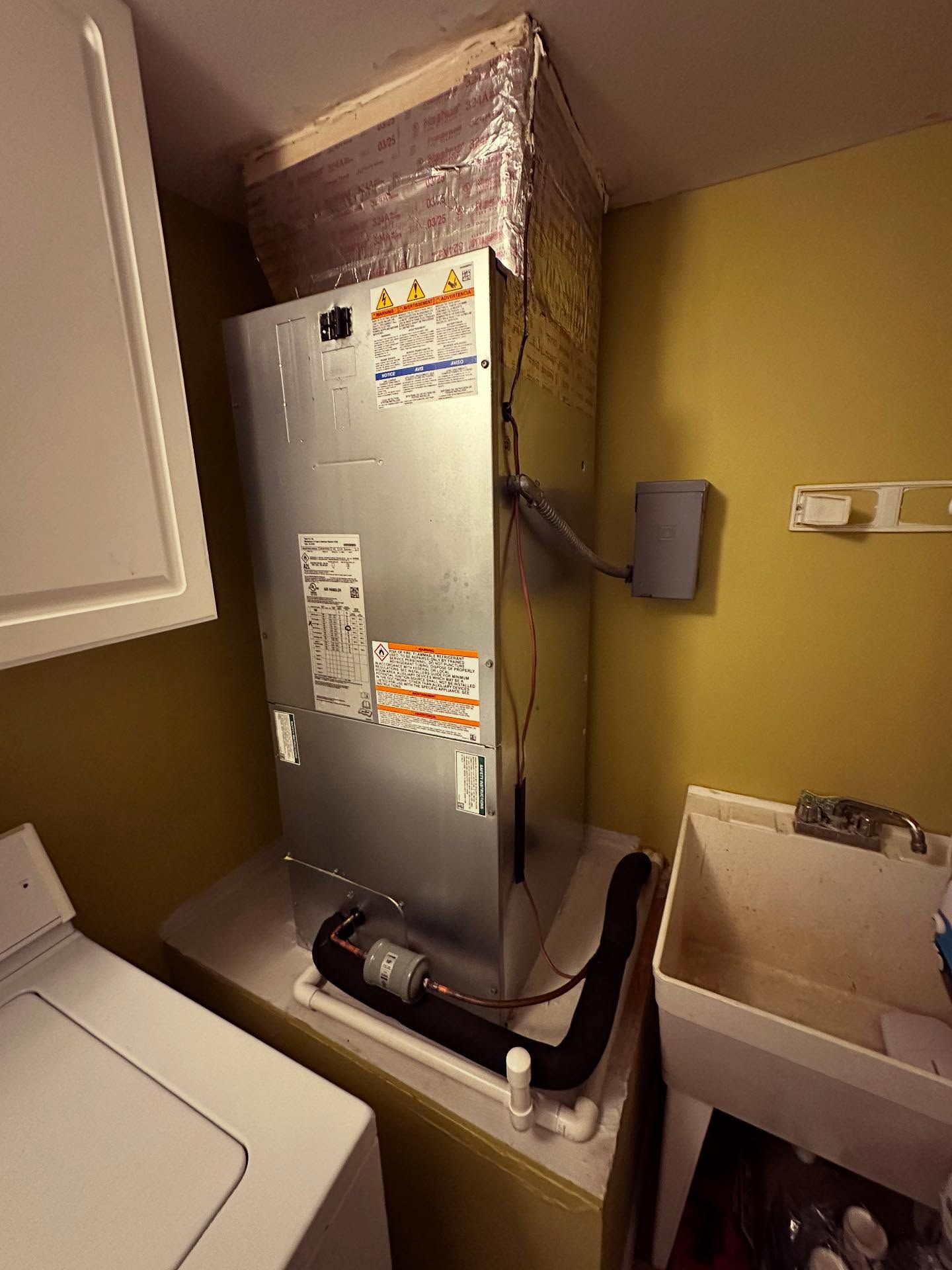 A tall, silver HVAC unit installed in a laundry room, positioned between a washing machine, sink, and cabinet.