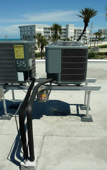 Two rooftop air conditioning units with black pipes, next to a building and the ocean, under a blue sky.