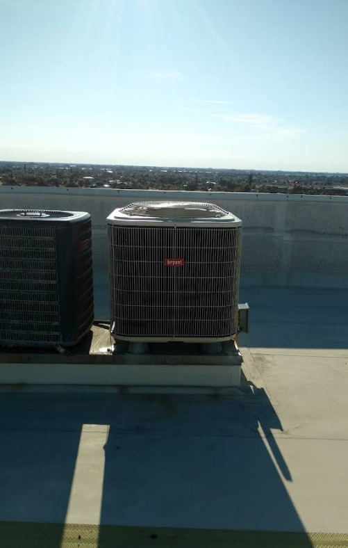 Two rooftop air conditioning units under a sunny sky, with a city visible in the background.