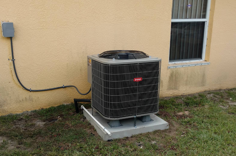 An air conditioning unit next to a house. The unit is black and gray, with a power box on the wall above it.