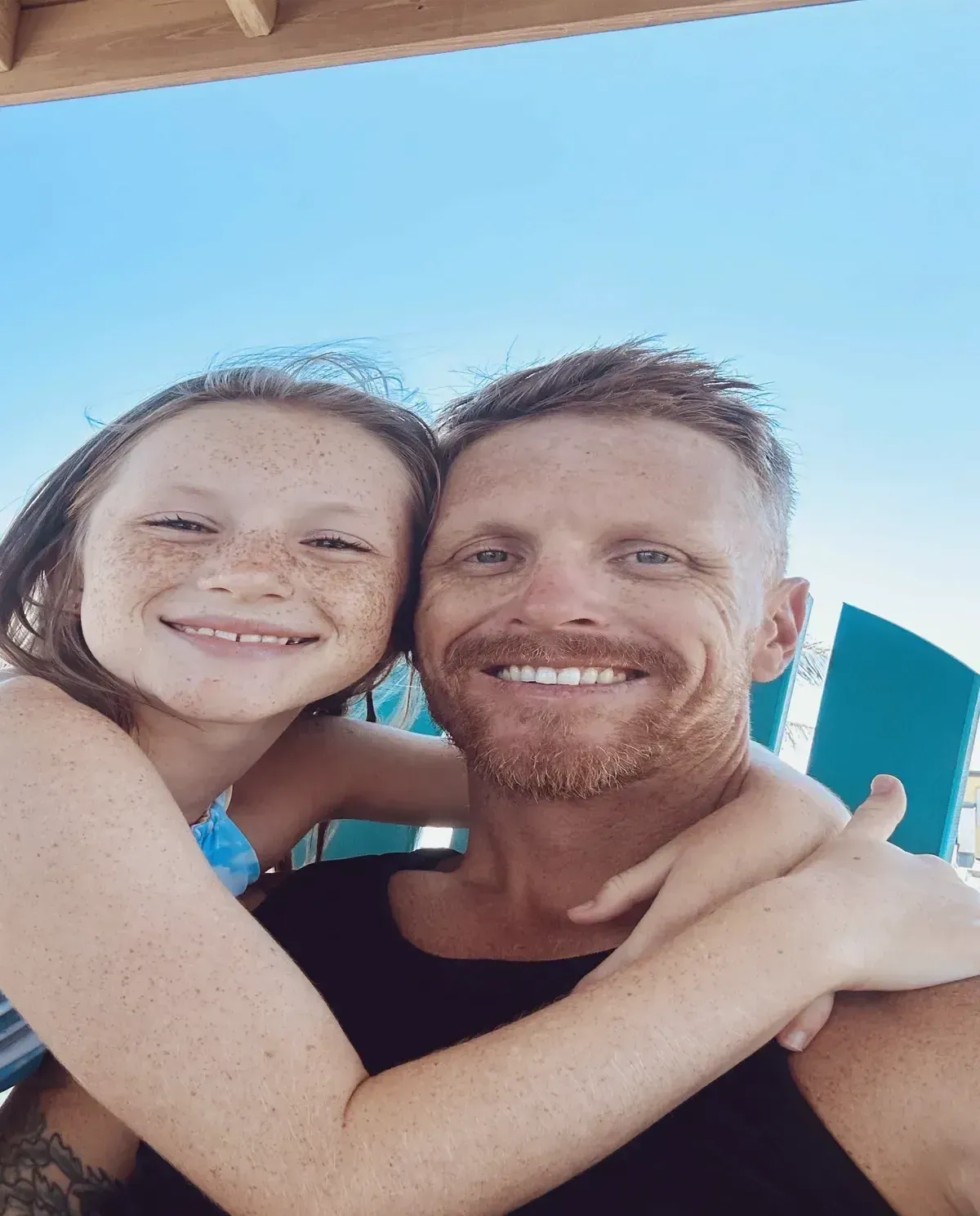 Father and daughter smiling, embracing outdoors. The man has reddish hair and beard, and the girl has freckles.