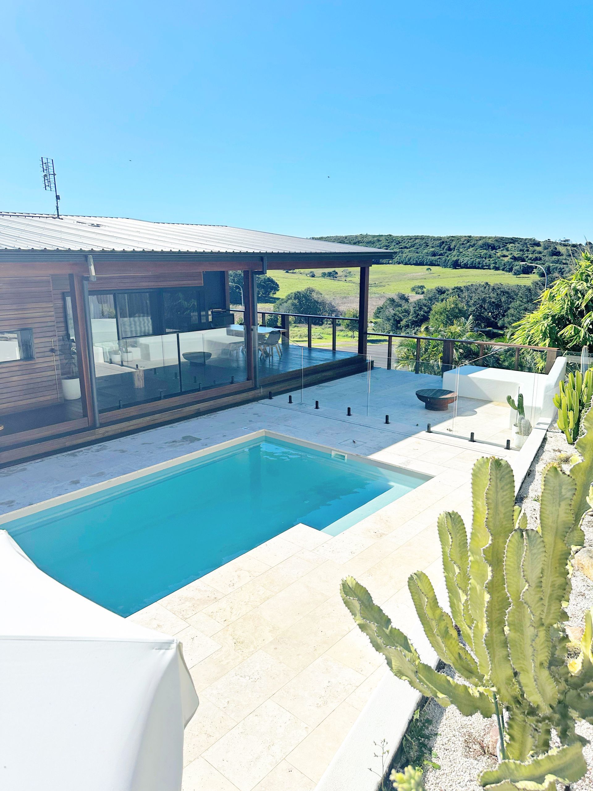 Swimming pool next to a wooden building with glass walls, overlooking green hills under a blue sky — Evoke Pools In Lennox Head, NSW