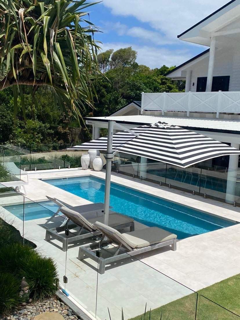 A white two-story house with a pool and striped umbrella. Lounge chairs sit poolside on a sunny day — Evoke Pools In Lennox Head, NSW