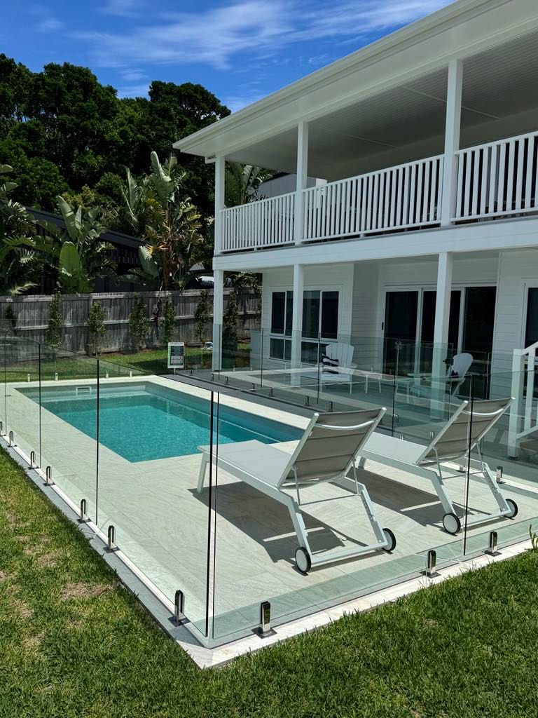 Two-story white house with a pool. Lounge chairs sit by the clear glass fence surrounding the pool. Lush green lawn — Evoke Pools In Lennox Head, NSW