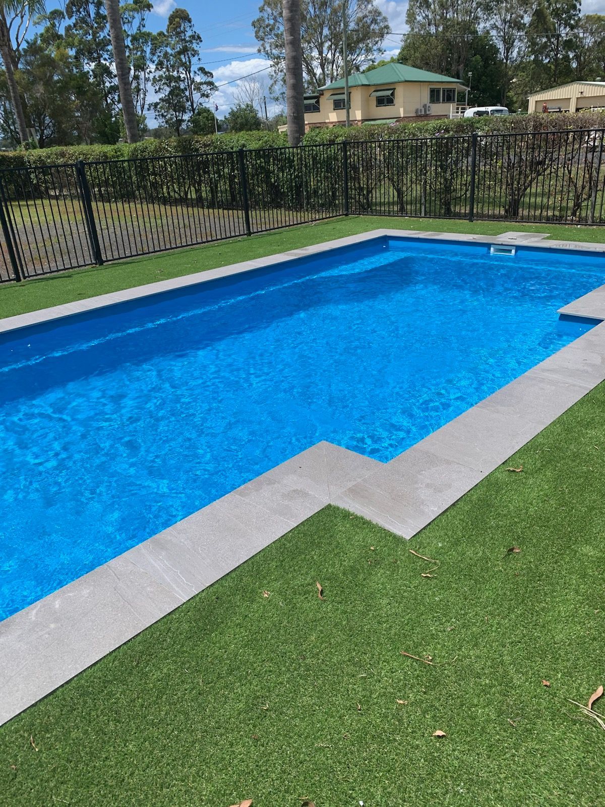 Blue pool surrounded by gray paving and green artificial turf, black fence in background, sunny day — Evoke Pools In Lennox Head, NSW