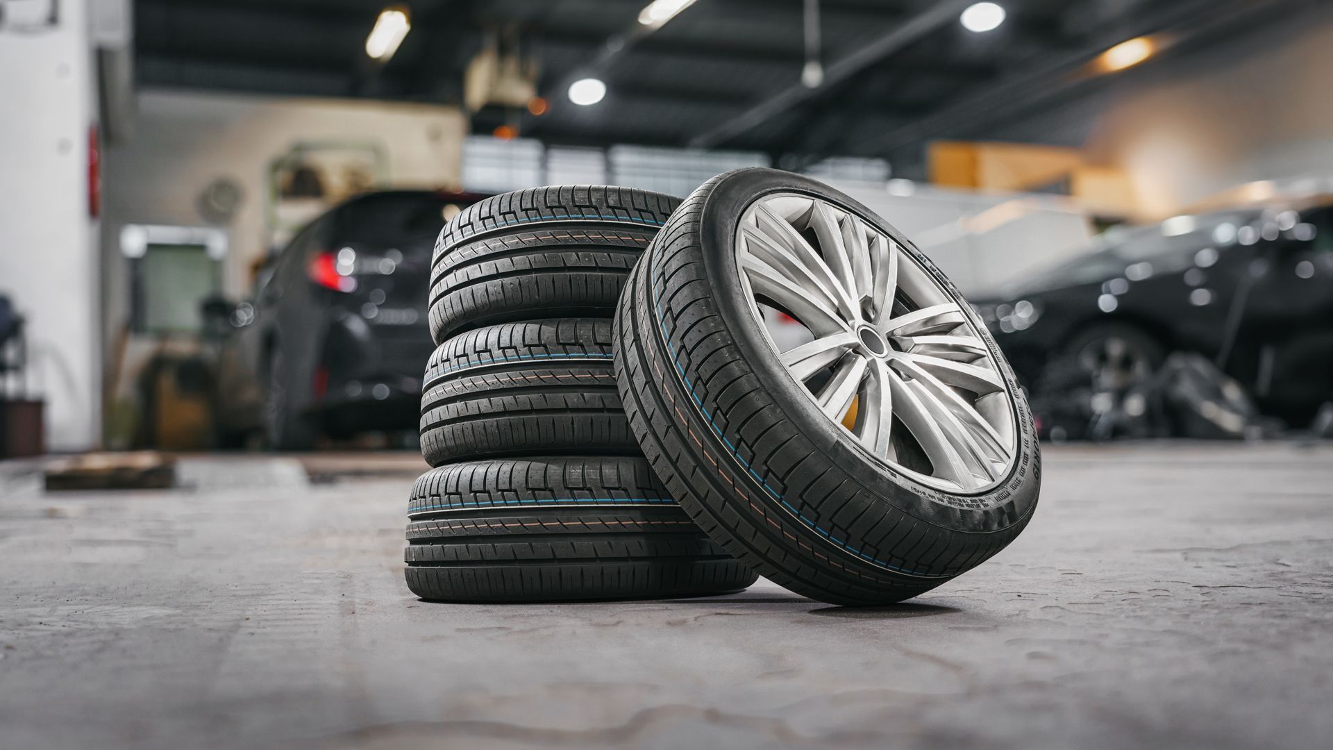 Four new tires stacked, next to a mounted wheel, in a garage with cars.