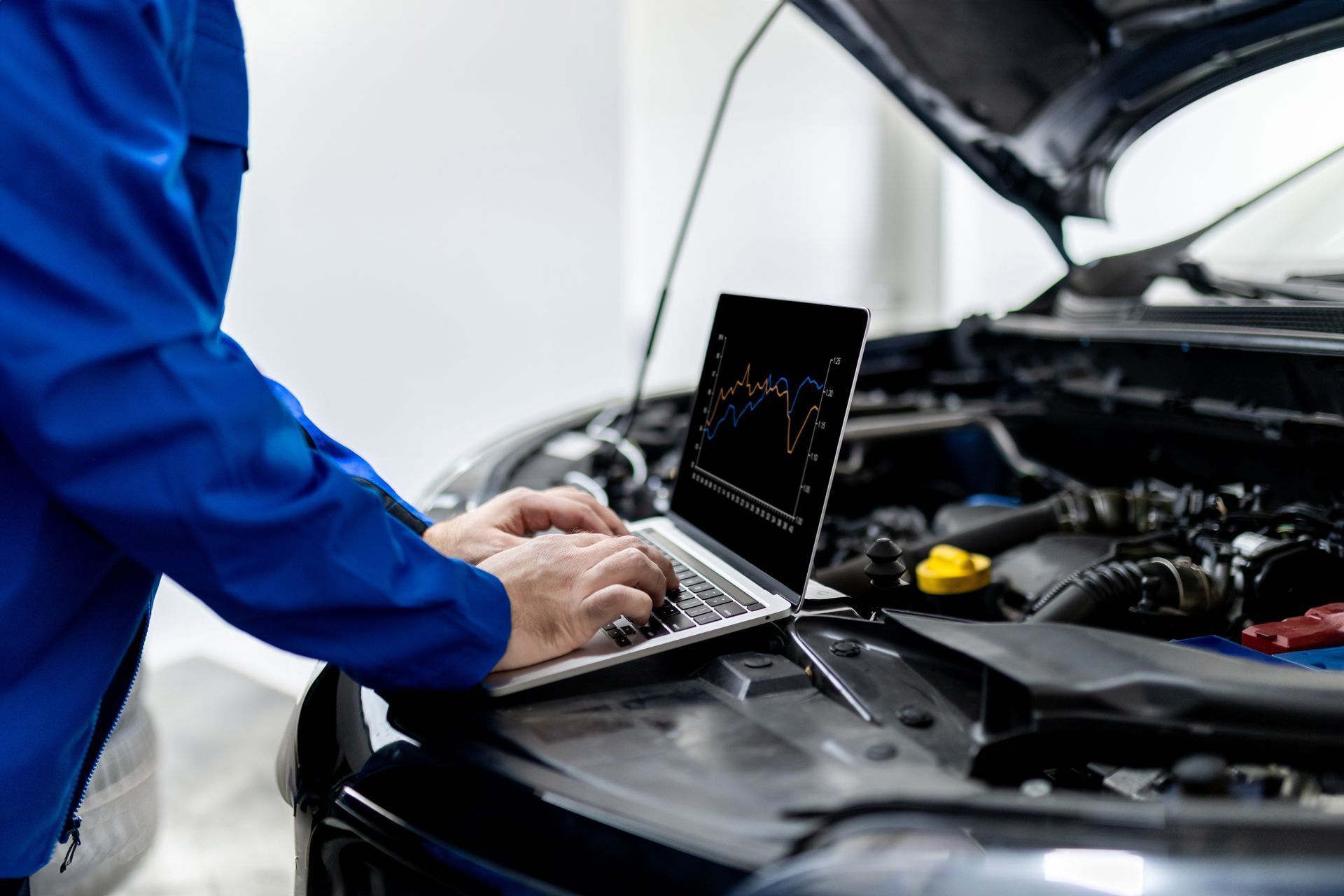 Mechanic using laptop to diagnose a car engine with hood open.