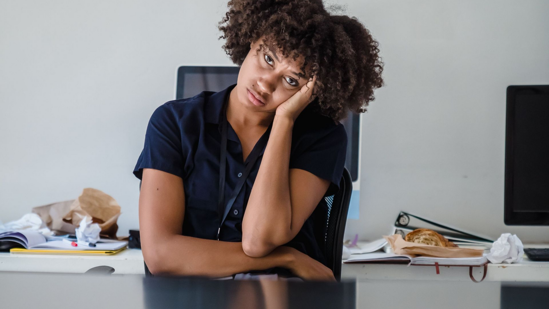 Person sitting at a desk, leaning on one hand with a tired expression, papers and office items in the background