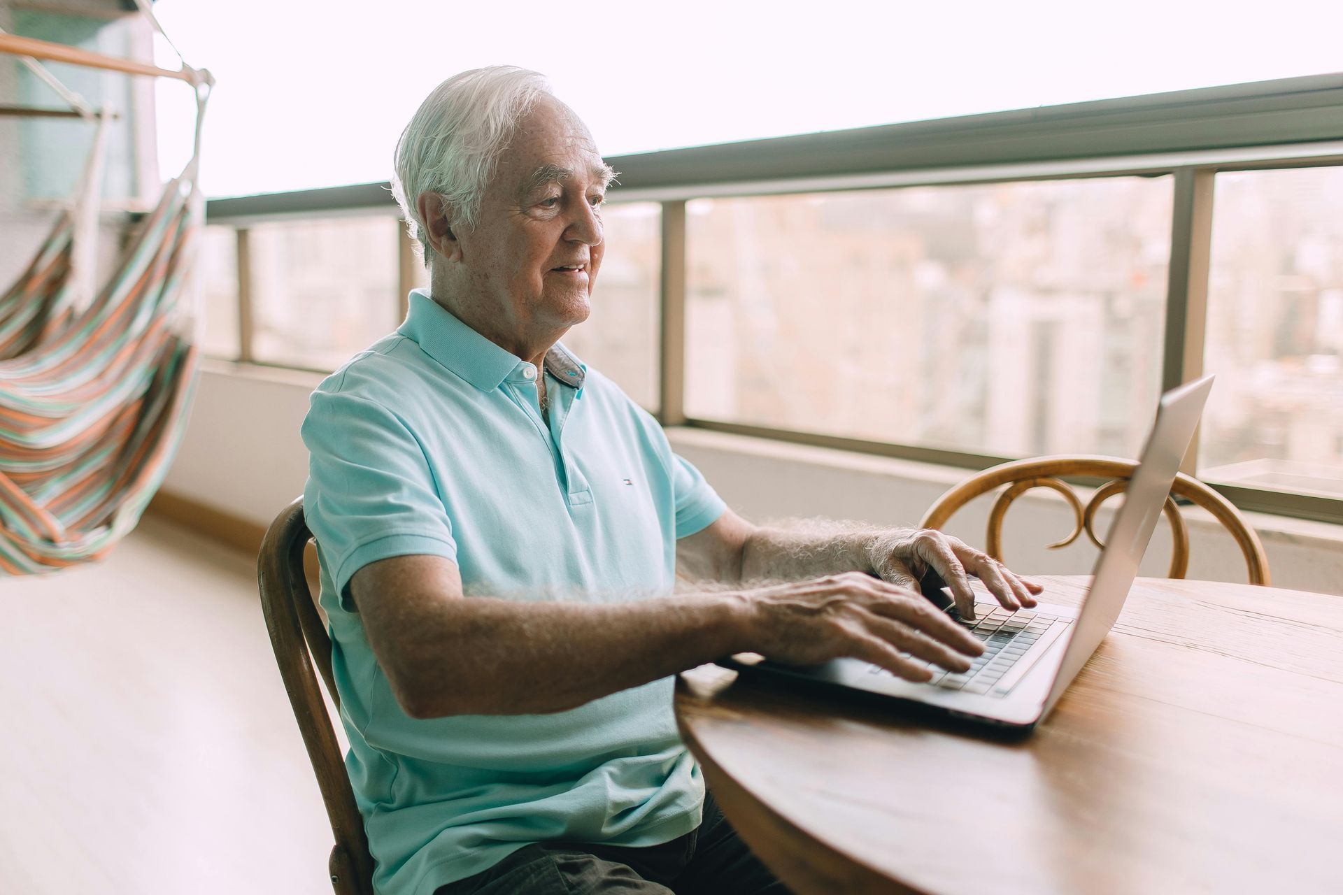 Older man wearing a light blue shirt, smiles as he types on a laptop at a round table.