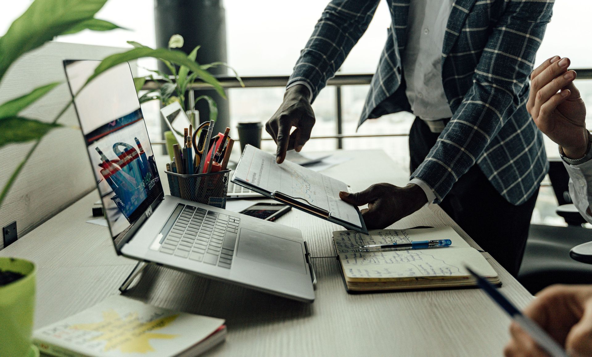 People collaborating at a desk with a laptop, documents, pencils, and a plant in a bright office