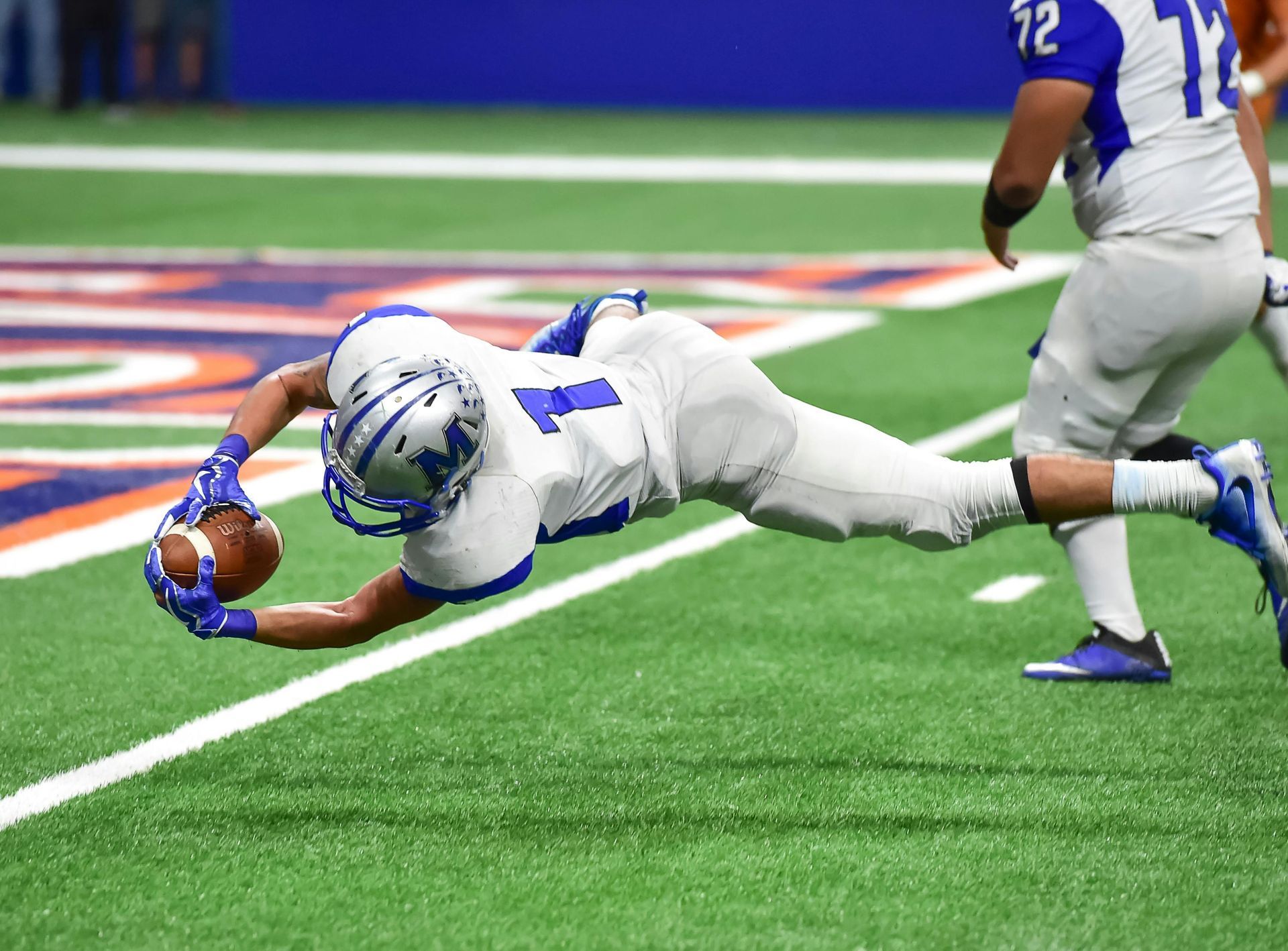 Football player in blue and white uniform diving for a catch on a green field.