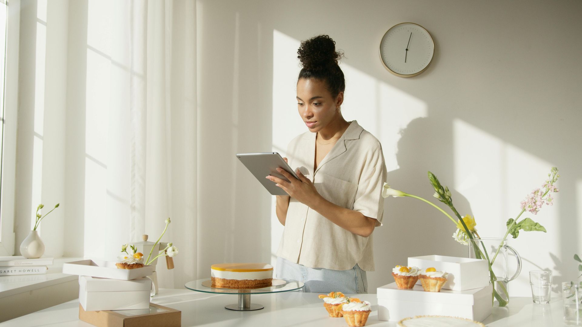 Woman in a bright kitchen looks at a tablet near a cake and cupcakes. Sunlight streams through a window.
