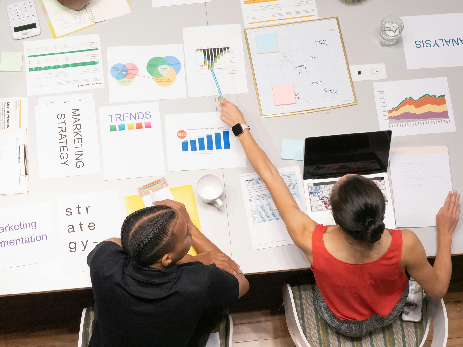 Two people reviewing colorful charts and notes on a table with a laptop in a bright office