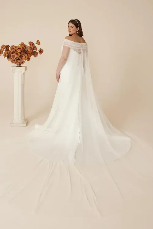 Bride in white off-the-shoulder wedding dress with flowing cape, standing in a studio, next to flowers.