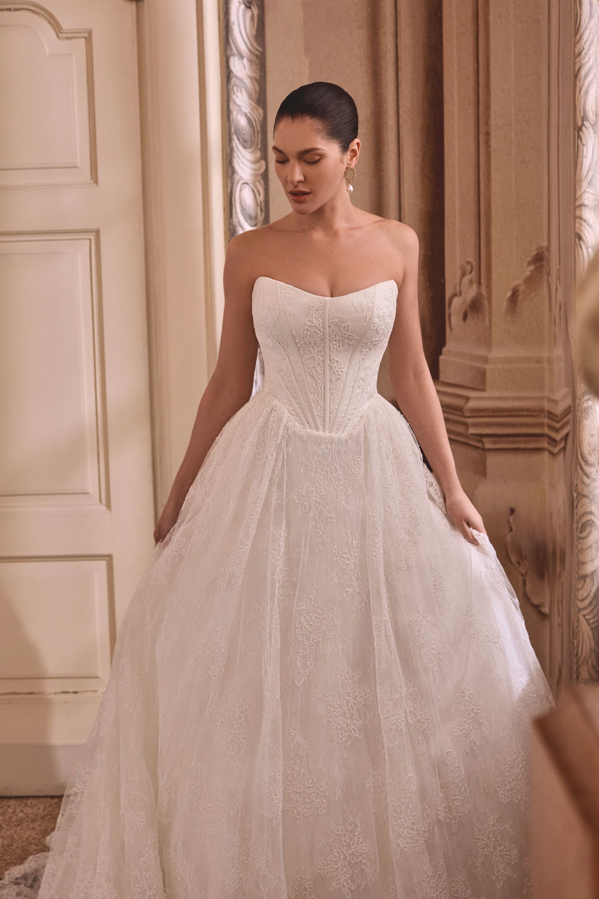 Woman in white wedding dress with open lace back and train, standing in a bright room.