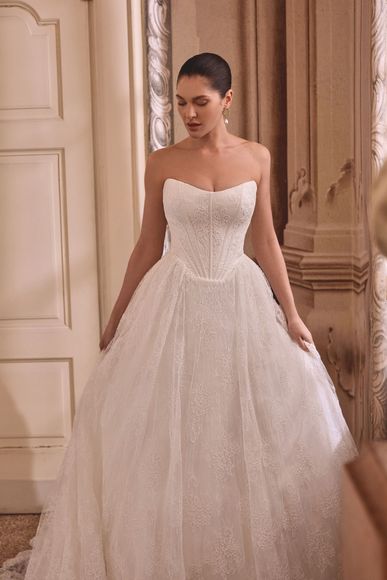 Woman in white wedding dress with open lace back and train, standing in a bright room.