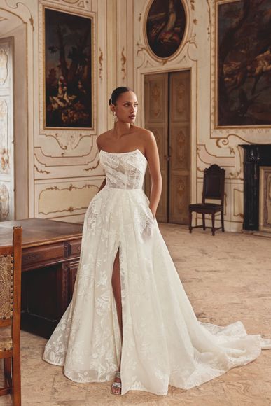 Woman in white wedding dress with open lace back and train, standing in a bright room.