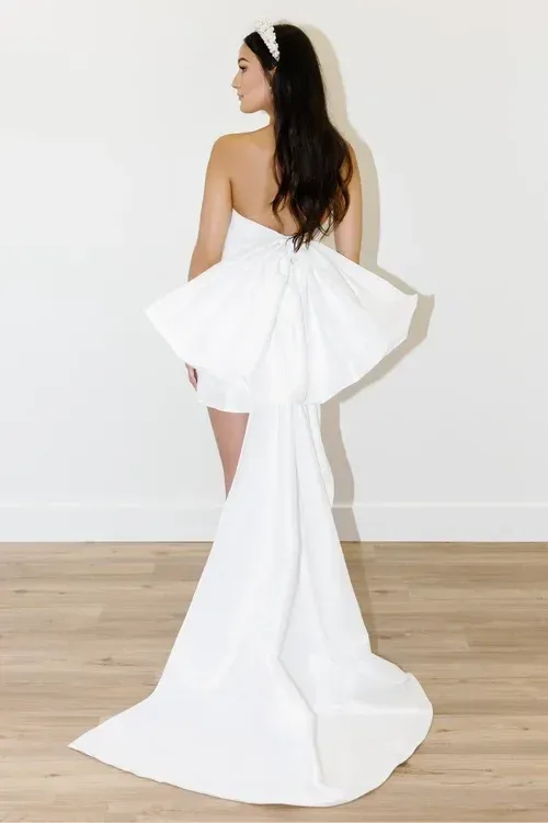 Woman in white strapless dress with large bow back and train, wearing a headband, facing away from camera.